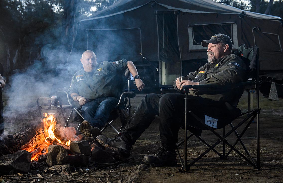 Two men sit next to an open fire in the campsite 