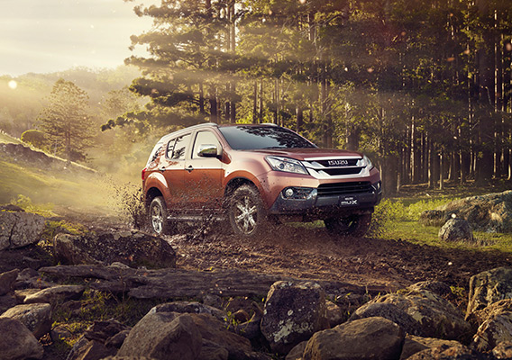 Isuzu MU-X drives on rocky track through forest with light beams behind
