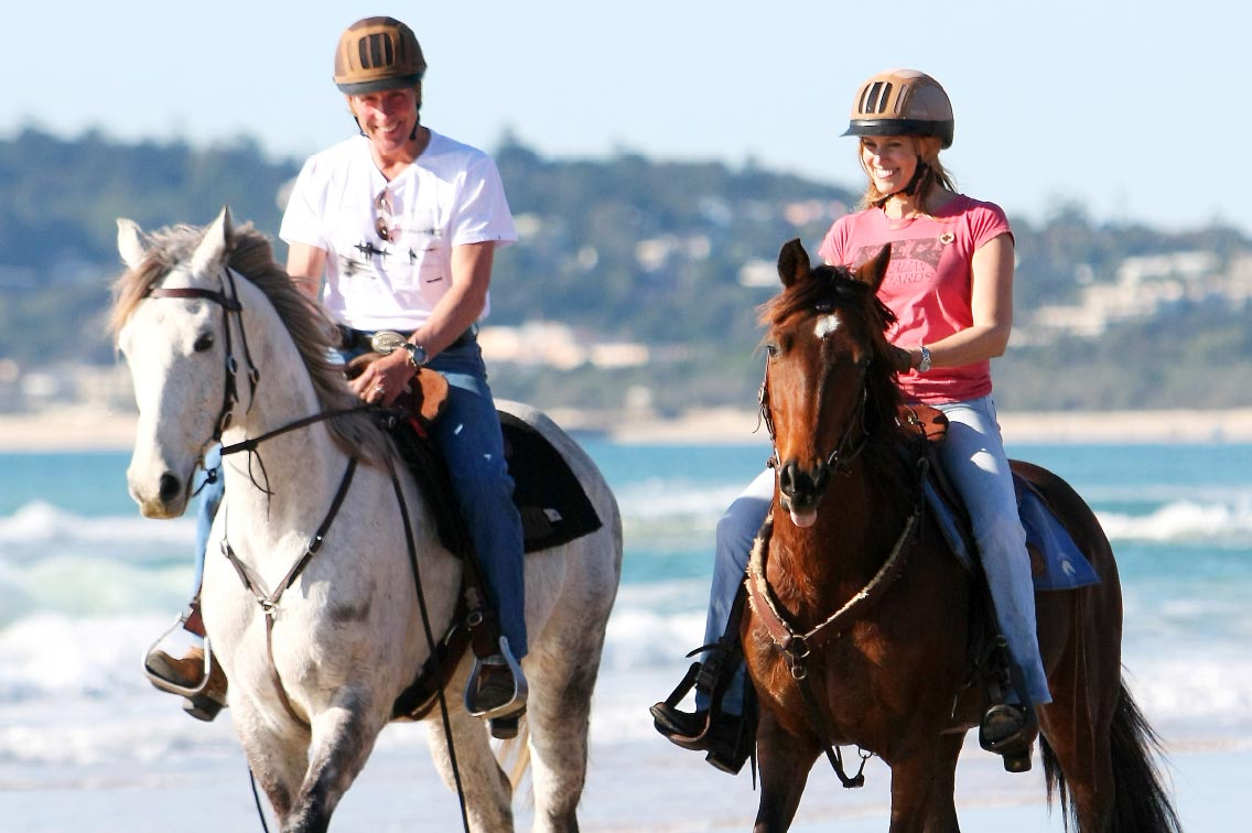 Two people ride horses along beach