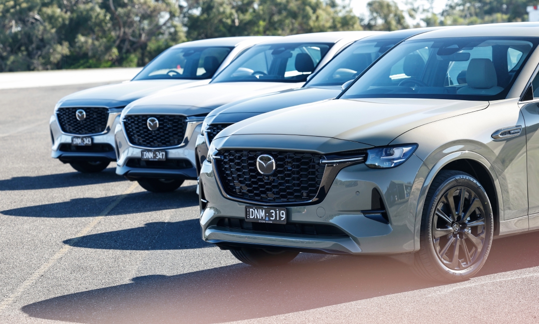 Four 2025 Mazda CX-60 vehicles parked in a row on an outdoor surface, angled toward the camera, with greenery and trees in the background.
