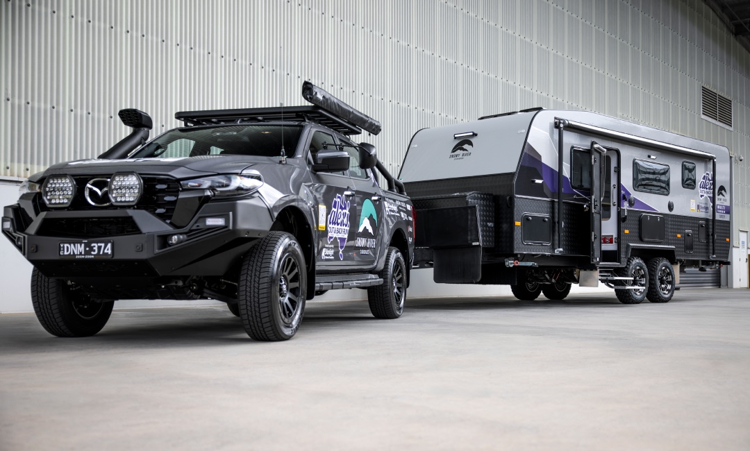 Mazda BT-50 ute towing a large camper, branded with Alex Barbas sponsor logos, parked in front of a warehouse — showcasing rugged adventure gear and caravan lifestyle branding.