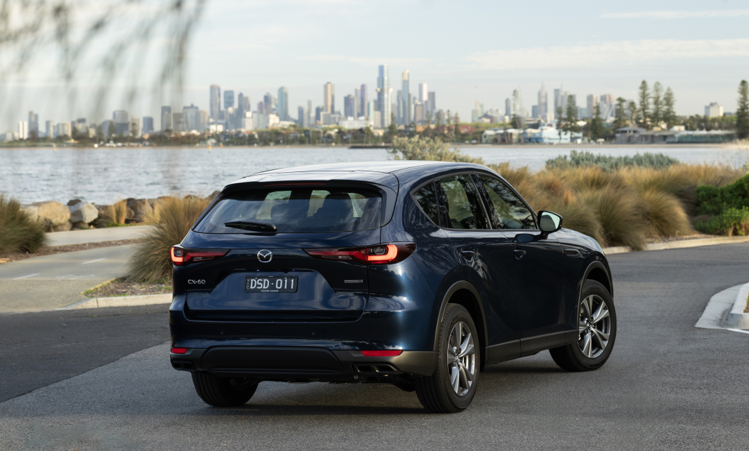 Rear view of a blue Mazda CX-60 G25 SUV parked near a waterfront with Melbourne city skyline in the background.