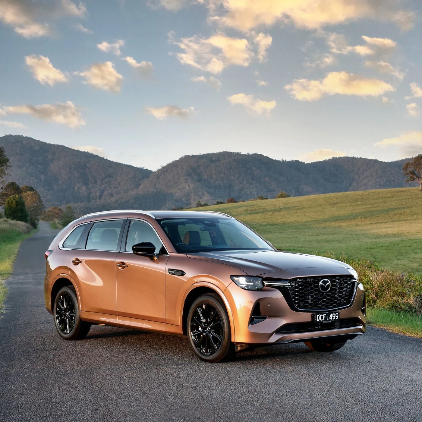Melting Copper Mazda CX-80 SUV parked on a country road with green fields and forested hills in the background, featuring sleek design, black alloy wheels, and LED headlights.