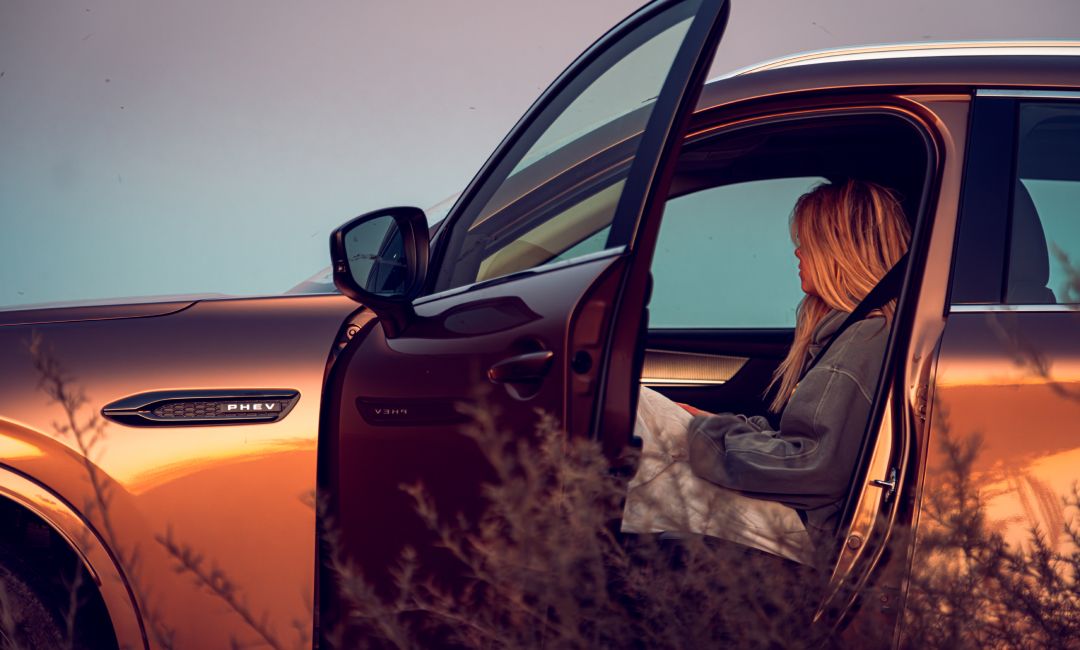 Mazda CX-80 in Melting Copper parked on an outback track in Western Australia during wildflower road trip, passenger seated inside with door open, enjoying the rugged landscape at dusk.