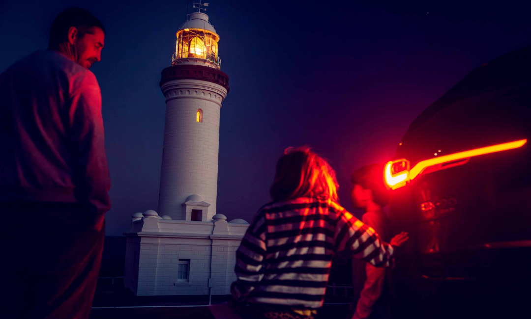 Norah Head Lighthouse lit at night as a family stands by the glowing taillight of the Mazda CX-90