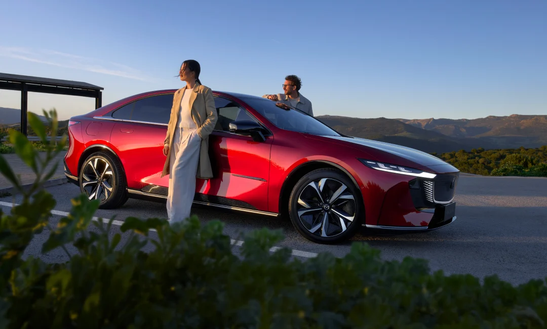 Mazda 6e electric vehicle in red parked at a scenic mountain lookout during sunset, with two people standing beside the EV in a lifestyle outdoor setting.