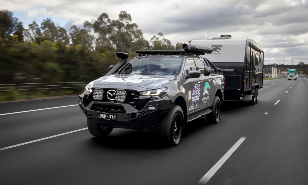 Mazda BT-50 ute towing a large camper trailer on a highway, branded with Alex Barbas sponsor logos, surrounded by trees and passing vehicles — highlighting rugged travel and caravan lifestyle