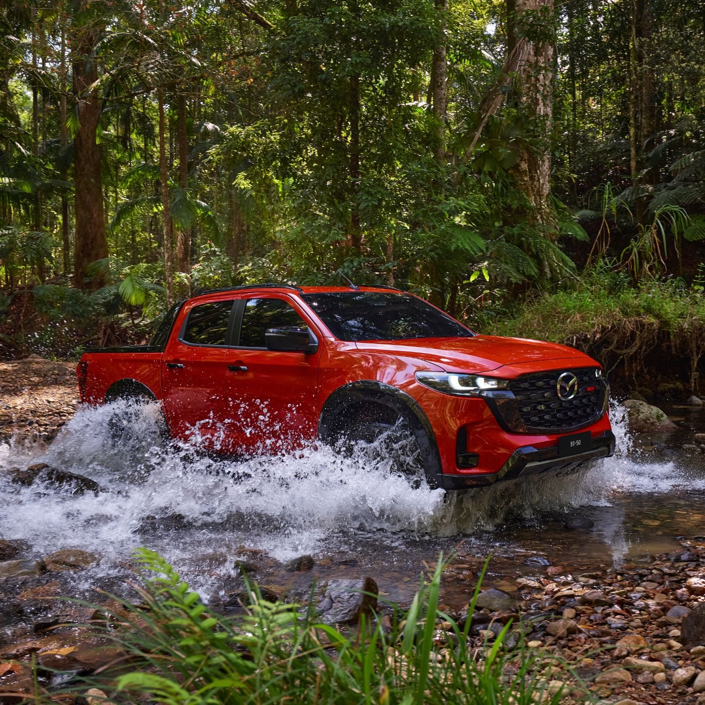 All-New 2025 Red Earth Mazda BT-50 ute driving through water, showcasing off-road capabilities in a dense tropical forest.