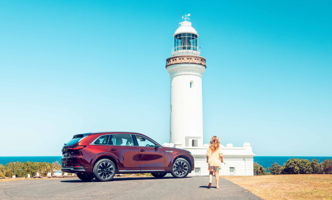  Mazda CX-90 Azami parked beside Norah Head Lighthouse as a girl walks toward the tower